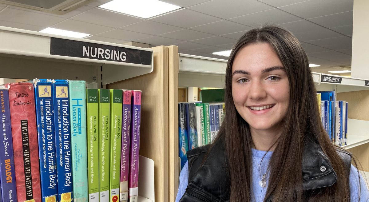 Nadia Hall, female with long dark hair wearing a blue top with black body warmer pictured in library with nursing books in background.